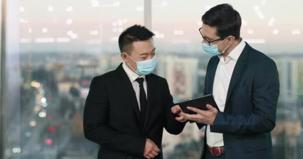 两个穿着雅致西服的混血工人在谈论发展商业 戴口罩的男性在使用平板电脑时积极交流 Coronavirus流行病概念 — 图库视频影像