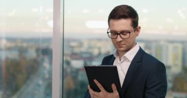 Ofisteki panorama penceresinin yanında duran gözlüklü, şık takım elbiseli yakışıklı beyaz iş adamı. Tablet bilgisayar ekranına tıklamakla meşgul. Teknoloji aygıtı ile çalışmak.