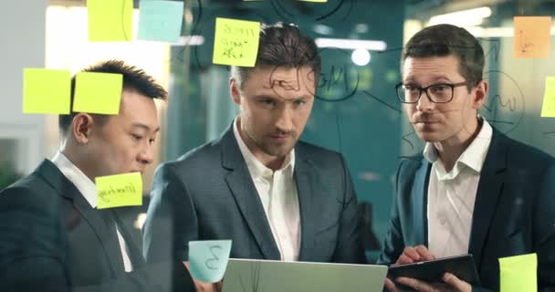 3人のリーダーのチームが戦略ビジネスプロジェクトについて決定します 近代的なオフィスで会っている深刻な人々 ガラス板の後ろに立ち グラフを検討する 技術を使って — ストック動画