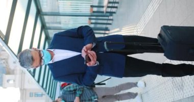 Takım elbiseli yakışıklı adam seyahati için yola çıkmayı beklerken telefonu kullanıyor. Koronavirüs salgını sırasında koruyucu maske takan insanlar. İstasyondaki turistlere dikey görüş. Turizm konsepti.