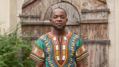 Orta yaşlı Afrikalı adam kameraya poz veriyor. Geleneksel kıyafet giymiş bir erkek gülümsüyor ve düzgün görünüyor. Çapraz kollu süslü elbiseli tatmin olmuş bir adam. Çok ırklı bir adam kameraya göz kırpıyor.
