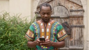 Süslü kostümlü melez bir adam. Geleneksel parlak kıyafetler giymiş Afrikalı bir adam. Çok kültürlü bir adam telefonda gülümsüyor ve mesajlaşıyor. Afrika kıyafeti giymiş bir erkek. Elinde bir alet var..