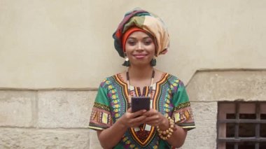 Afrika görünümlü güzel bir kadın kameraya bakıyor. Kafasında Afrika ambalajı olan kadın gülümsüyor. Geleneksel mücevher işinde çok ırklı bir kadın telefon tutuyor. Afrikalı bir kadın..