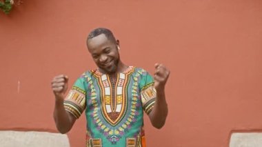 Orta yaşlı Afrikalı adam kırmızı duvarda dans ediyor. Çok kültürlü bir adam eğleniyor ve parmaklarını şaklatıyor. Geleneksel süslemeli gömlekli bir erkek. Adam kablosuz kulaklıkla müzik dinliyor..