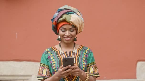 伝統的な服を着たアフリカ人女性の肖像画 若い笑顔のインド人女性がターバンで 赤い壁を背景にジュエリーを持つ多文化の女性 携帯電話を手にした多民族女性 — ストック動画