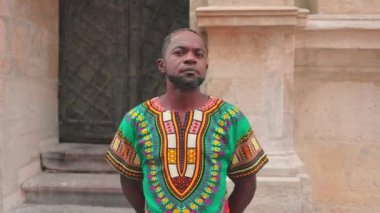 Renkli gömlekli Afrikalı adam başını kameraya çeviriyor. Kamera için geleneksel süslenmiş pozlar veren bir erkek. Afrika görünümlü çok kültürlü bir adam ellerini arkaya koydu..