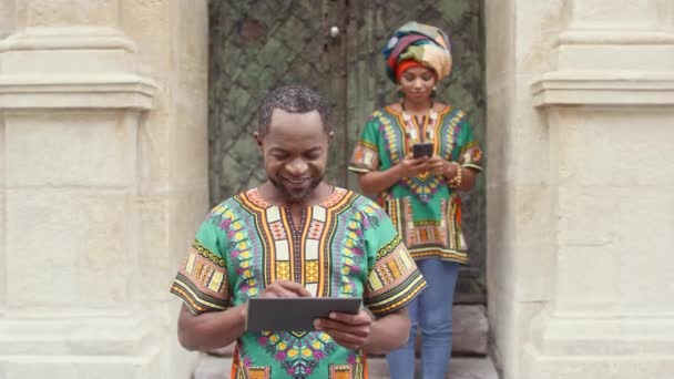 ドアの後ろに明るいアフリカの服の男と女 アフリカ人男性の装飾シャツの前にタブレットを保持する スマートフォンを背景にしたアフリカ人女性 — ストック動画