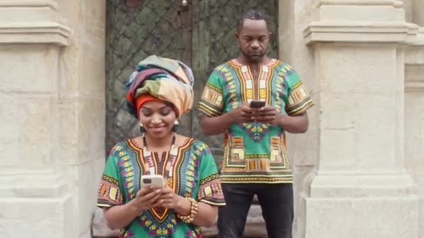 アフリカの明るい服のドアの背景にポーズをカップル笑みを浮かべて 女性の前で伝統的なターバンで シャツの後ろに男 多文化カップルでお祝いアフリカの服を携帯電話を保持 — ストック動画
