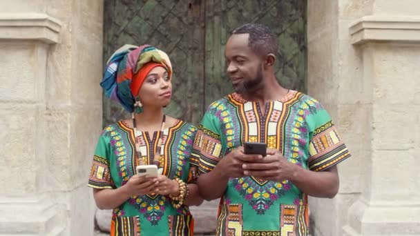 中年のアフリカのカップルのカメラでポーズ以外のお互いを見ています 多民族のカップルがスマートフォンを持っています メイクやスカーフを持つ女性 伝統衣装を着た多文化の人々 — ストック動画