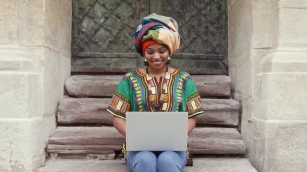 穿着鲜艳衣服头饰的成熟的非洲女人 笑着多文化女人坐在楼梯上和笔记本电脑上工作 穿着传统衬衫和头巾看笔记本电脑的非洲妇女 — 图库视频影像