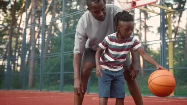少年は公園の屋外裁判所でバスケットボールボールを跳ねる アフリカ系アメリカ人の父親の世話でバスケットボールを練習している小さな子供 スポーツレジャーや週末 家族の時間だ バスケットボールゲーム — ストック動画