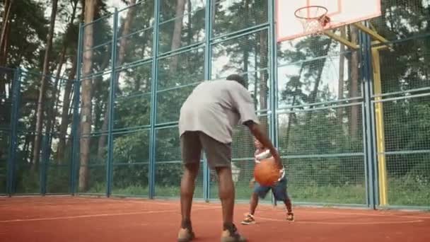 アフリカ系アメリカ人のかわいい男の子が遊び場の外で大人の父親とバスケットボールをします 大人の男が跳ねてボールを投げ込む 父と息子はスポーツゲームをプレイし 楽しみを持って — ストック動画