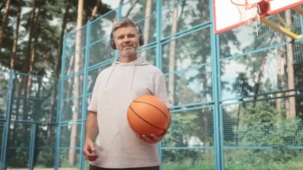 Kulaklıklı Yetişkin Bir Adam Basketbol Sahasında Durup Kameraya Gülümseyerek Bakıyor — Stok video
