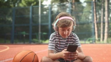 Basketbol sahasında oturan ve açık havada akıllı telefon kullanan sevimli ergen çocuk. Kulaklıklı küçük bir çocuğun açık havada müzik dinlediği portresi. Boş zaman harcamak.