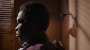 Ofisinin koltuğunda oturan takım elbiseli Afro-Amerikan bir adamın yan görüntüsü. Gölge odasında biriyle konuşuyordu. Arka planda parlak ışık ve pencerenin perdeleri ile görünür lamba.