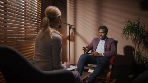 影の部屋で患者をカウンセリングする女性心理学者のリアビュー 彼のセラピストに何かを伝える優雅なスーツで成功したビジネスマン 椅子に座っている間にコミュニケーションをとる多民族 — ストック動画