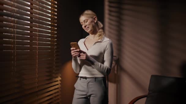 シャドウオフィスのスマートフォンにメッセージを入力する美しい白人女性 ポジティブな女性は 盲目の窓の近くの誰かとチャットします 良いメッセージを読んだ後に良い気持ちを持つこと — ストック動画