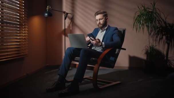 アームチェアに座っている間 スマートフォンで誰かとチャットするエレガントなスーツの良い見栄えの白人男性 シャドールームのリモート通信中にノートパソコンを使用する若いビジネスマン — ストック動画