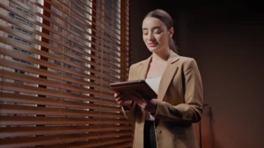 Kafkasyalı genç bir kadının tablet cihazını kullanarak yaklaşırken büyük pencerenin yanında ahşap perdelerle dikiliyordu. İnternet sitelerinde video izlerken kulaklık takmak. Teknoloji kavramı.
