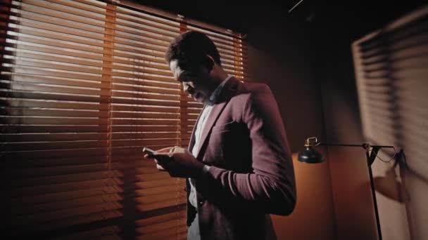 アフリカ系アメリカ人の若い男性が 木製のブラインドで隣の窓に立っている間 スマートフォンを使用しています ウェブサイトやソーシャルメディアをスクロールする ダークオフィスで一人で過ごす時間 — ストック動画