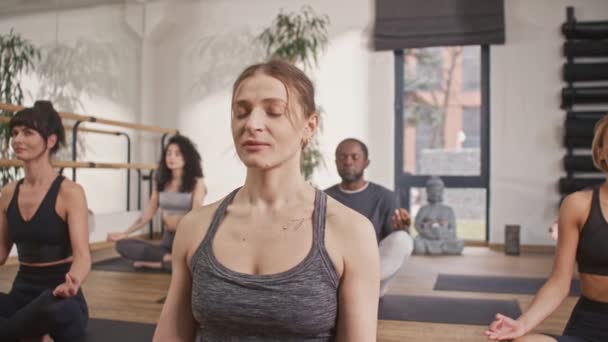 ヨガマットで瞑想するフィットネス服を着た混合スポーティな人 目を閉じたロッツポジションで快適に座っています 深呼吸を行い 内側の精神的なエネルギーに集中する — ストック動画