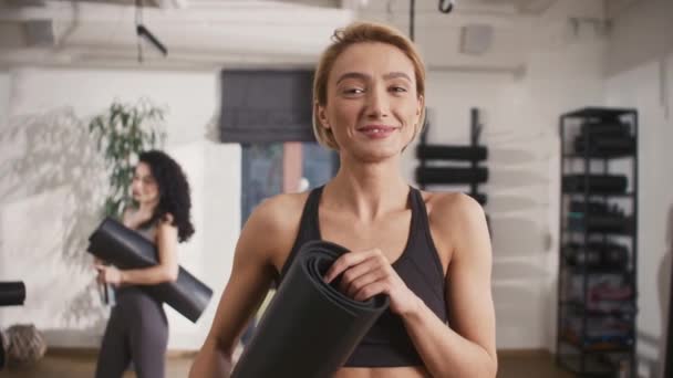 白种人女教练的画像 拿着健身垫 在镜头前欢快地微笑 在背景下 快乐的女人经过艰苦的训练后 彼此积极地交谈 — 图库视频影像