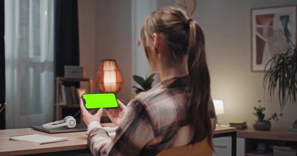 坐在家里看绿色手机屏幕的女人 女性聚焦并观看彩色键屏 女人坐在桌旁拿着智能手机女人坐在满是文具的桌旁 — 图库视频影像