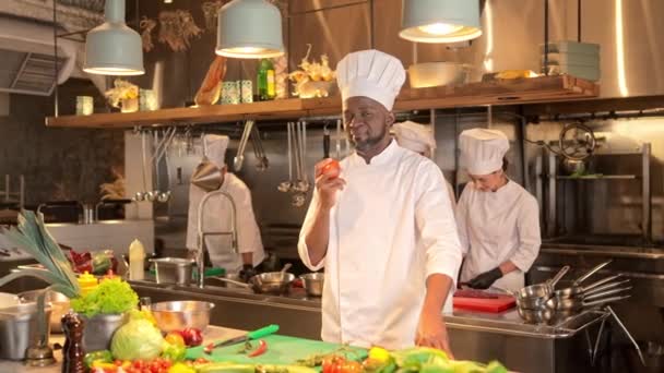 アフリカ系アメリカ人男性シェフが立ってジャグジーのトマト 多文化料理は レストランのキッチンの背景に立って カメラと笑顔を見ています 一緒に働くクックのプロキシアルバム — ストック動画