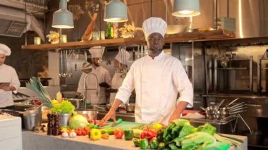 Afro-Amerikalı aşçı kollarını kavuşturup kameraya bakıyor ve gülümsüyor. Restoran mutfağının arka planında çalışan tatmin olmuş bir aşçı. Çok kültürlü aşçılardan oluşan profesyonel bir ekip.