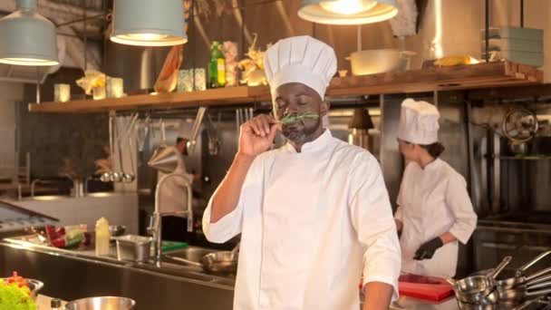 アフリカ系アメリカ人の男性シェフは レストランのプロのキッチンに立っており 香りの良いハーブスパイスの香りがします 多文化的な笑顔の料理は料理の準備のための材料を選びます — ストック動画