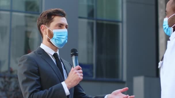 保護マスクを着用している男性ジャーナリスト特派員にインタビューを行う多民族代表医師 建物の近くに住んでいる パンデミックコンセプト Covid 19について 医学と医療について — ストック動画