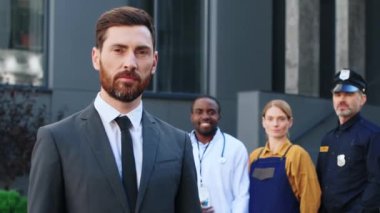 İyi giyimli bir ofis çalışanının portresi. Farklı mesleklerden farklı insanlardan oluşan farklı bir ekiple dışarıda duruyor. İş adamı gülümsüyor ve modern ticari binanın dışındaki kameraya bakıyor..