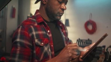Atölyedeki tamirci. Garajda duran ve elinde dijital tablet tutan yakışıklı adam. Afrika kökenli Amerikan oto tamircisi akıllı telefon kullanarak makine onarımı için araçlar sipariş eder..