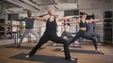 Orta yaşlı kadın koç, müşterilerine aerobik stüdyosunda yoga egzersizleri öğretiyor. Sportif çok ırklı insanlar spor paspasları üzerinde duruyor ve onların antrenör meditasyon hareketlerini takip ediyor.