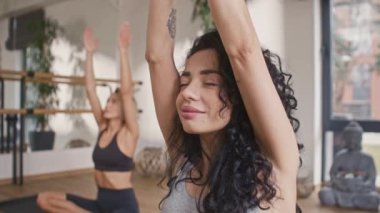 Spor bluz giyen güzel kıvırcık kızın portresi spor salonunda pilates yapıyor. Arka planda arkadaşı antrenman yapıyor. Kadınlar antrenörleriyle yoga yapıyor sakin ve rahatlatıcı müzik dinliyorlar..