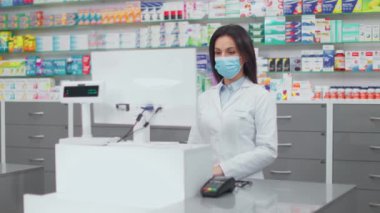 Bilgisayar ekranına bakan kadın eczacı. Kasada çalışan kadın eczacı. Vitrinin arka planında ilaç kullanan cübbeli ve maskeli bir kadın. Eczanedeki Kafkasyalı eczacı.