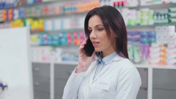 女药剂师看着电脑显示器 女药剂师通过电话与病人沟通 穿着白色长袍的女人通过电话交谈 用药物展示 药店里的白人药剂师 — 图库视频影像