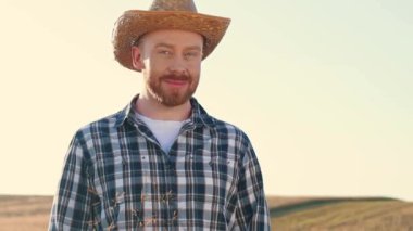 Yatay çekim. Arpa ya da buğday tarlasında duran erkek çiftçinin portresi. Hasır şapkalı ve kareli gömlekli bir adam. Tarım arazisinde duran neşeli çiftçi. Mutlu çiftçi dışarıda gülümsüyor.