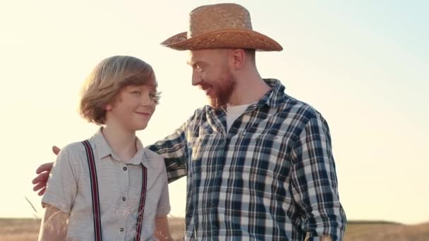侧向射击 快乐的男人抱着男孩 笑着站在田边的农民和男孩 男人穿着格子衬衫 戴着草帽抱着儿子 农业或耕作 快乐的男农和男童 — 图库视频影像