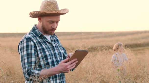 水平ショット 男の農夫がタブレットを持ち 息子と話している チェッカーされたシャツを着た男と ライに立っているわら帽子 収穫監監査を行っている農民の笑顔 フィールドに立っている陽気な農民 — ストック動画