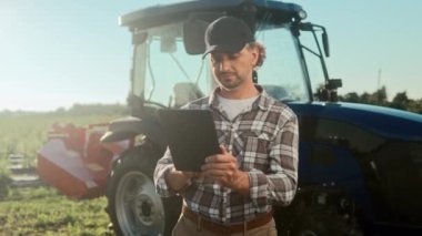 Erkek çiftçi tablet tutuyor ve hasat raporu veriyor. Traktör makinesinin arka planında duran tarım işçisi. Tablet kullanan kareli gömlekli ve şapkalı bir adam. Saban sürmeye hazırlanıyorum..