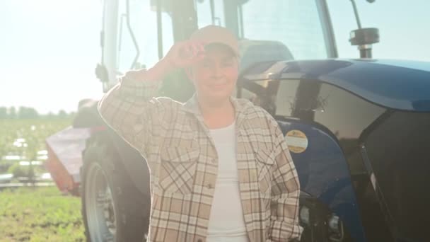 在阳光下站在田边的正高加索女人 笑着站在拖拉机后面的女农民 快乐的女人调整头上的帽子 穿着格子衬衫的女工 — 图库视频影像