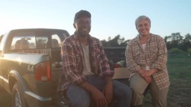 Kamyonetin bagajında oturan çok ırklı tarım işçileri. Tatmin olmuş erkek ve kadın ilk sebze mahsulünü topladı. Gülümseyen Afrikalı Amerikalı erkek ve beyaz kadın. Tarım mevsimi.