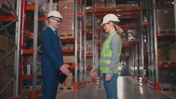 屋内で取引するコーカサス企業の労働者 反射ベストで女性請負業者に手を振る男性 倉庫に立っている保護ヘルメットの女性と男性 — ストック動画