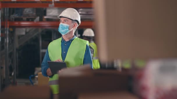 箱を持って棚の後ろにいる白人男性 倉庫で働いている男性 マスク 保護ヘルメット 黄色のベストで従業員を保管する タブレットを保持し 商品の量を計算する男性 — ストック動画