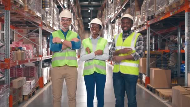 保護制服を着た多文化人が倉庫で互いを見つめあっている 3人の産業労働者が多層棚の背景で微笑んでいる 武器を持つストレージ従業員のチームが交差 — ストック動画