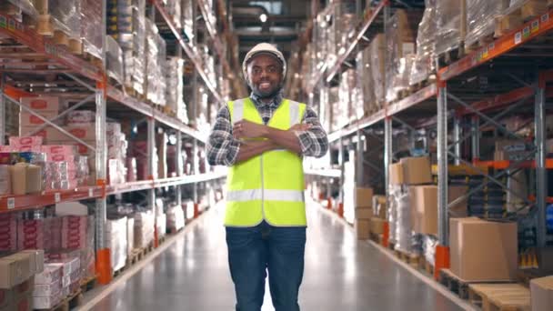 倉庫に立っているアフリカ系アメリカ人の笑顔 保護衣類とケーキの陽気な労働者 クロスアームでカメラを見ている男性請負業者 物流会社について 輸出のための商品 — ストック動画