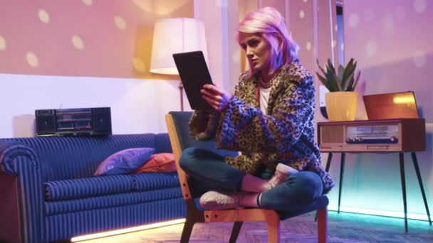 水平ショット 座ってタブレットを持っているヒップスターの女性 ディスコスタイルの客室でスタイリッシュな女性チリング ガジェットを持っている女性とインターネットでサーフィン レトロなヴィンテージルームでリラックスしたピンクの髪の女性 — ストック動画