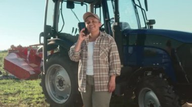 Kadın çiftçi telefonda neşeli bir şekilde konuşuyor. Kareli gömlek ve şapka giyen bir kadın. Traktör makinesinin arka planında duran kadın çiftçi. Tarım işçisi tarım arazisinde duruyor.