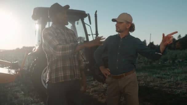 2人の農民が農業開発について話している 複数のフィールドワーカーが話し合っている トラクターマシンの背景にある農地に立っている男性 カウンティサイド農業 — ストック動画